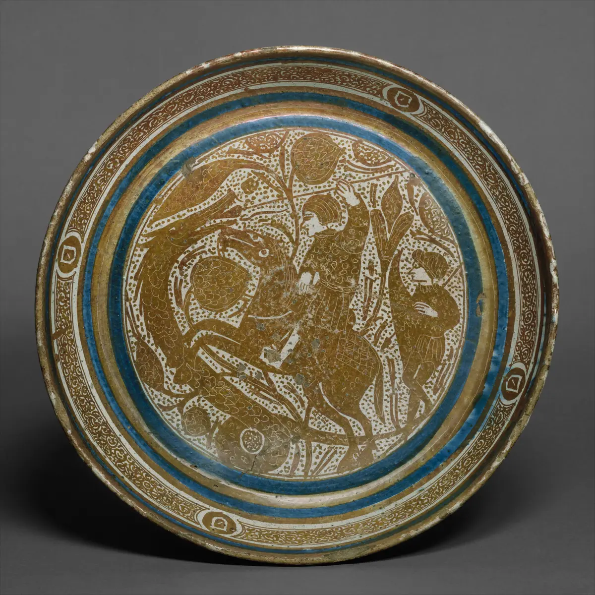 Cuenco de loza con reflejo dorado y bandas azules; en el centro, un jinete alancea a una serpiente entre motivos vegetales.