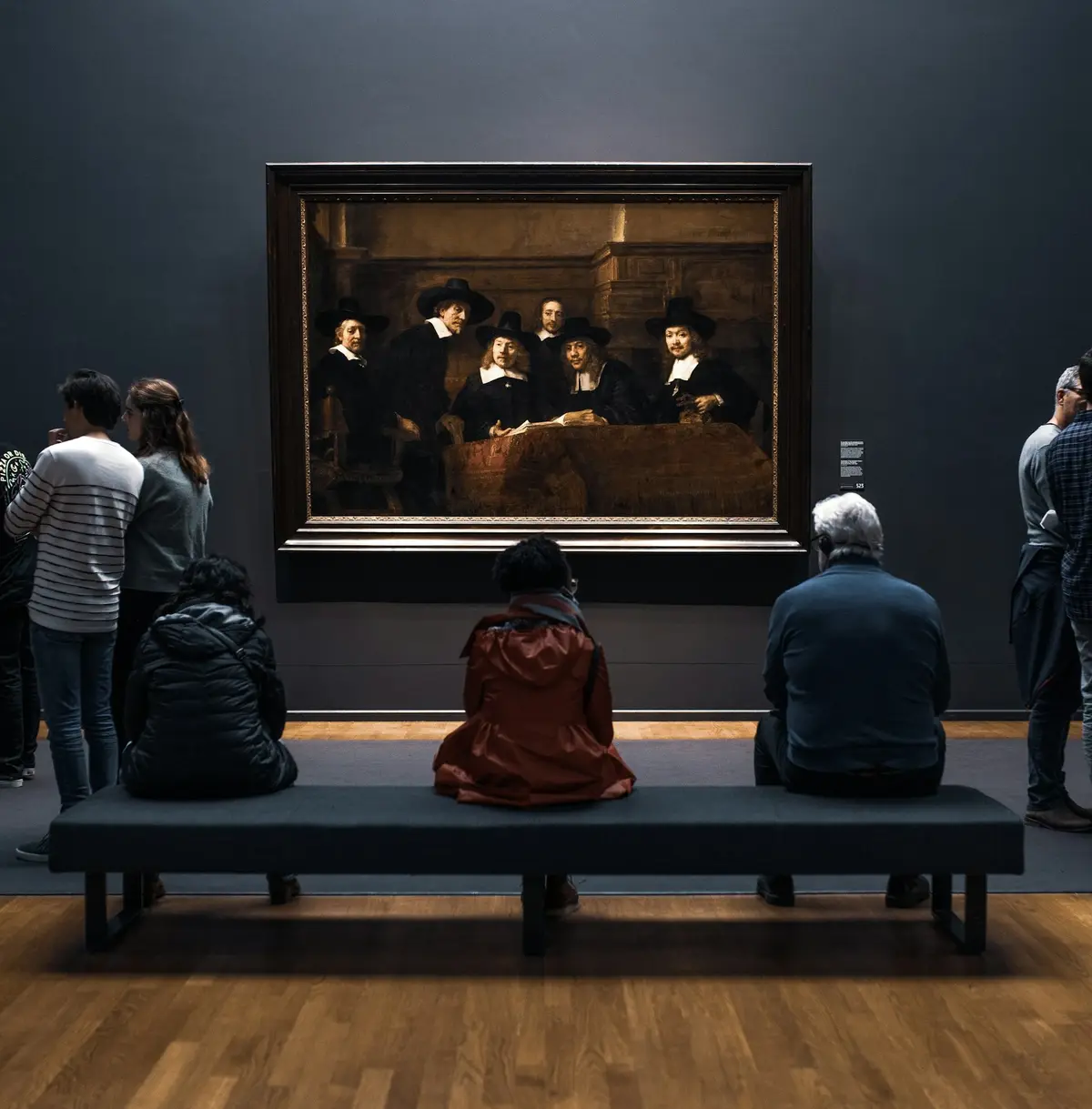 Visitantes en una sala del Rijksmuseum: tres personas sentadas en un banco y otras de pie contemplan un gran retrato de grupo de Rembrandt.